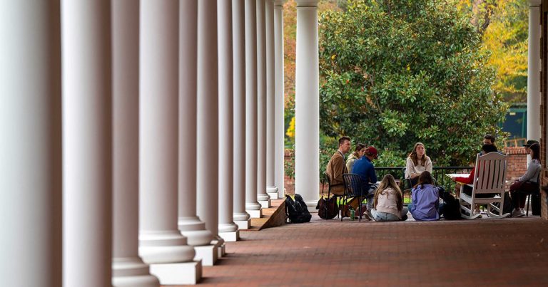 Ryan J. Johnson sits on a covered walkway, surrounded by students, with columns stretching to the left.