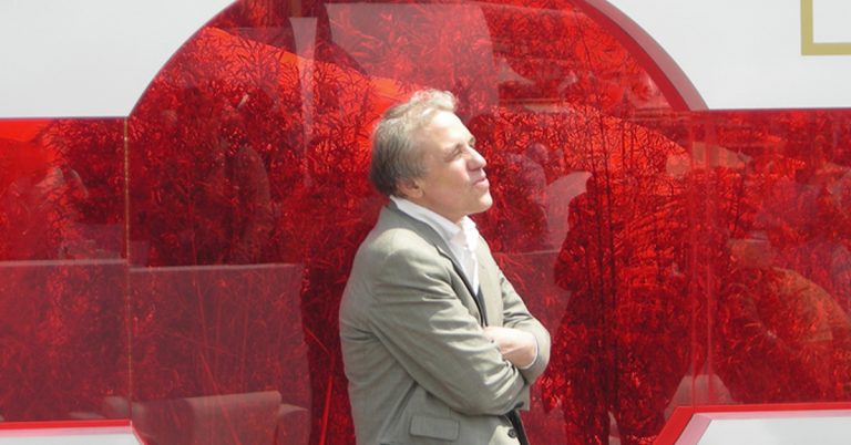 Abel Ferrara, a man with grey hair and dressed in a light grey suit, stands with his arms crossed in front of a red circle.