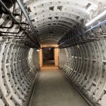 Looking down a round tunnel with strip lights and a small doorway at the far end.