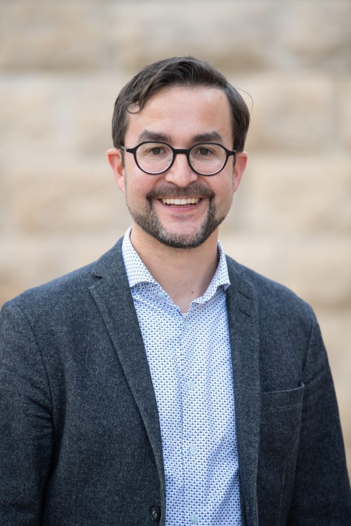 A photograph of Simon Fuchs smiling at the camera. He wears glasses, a shirt and a blazer.