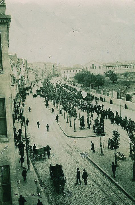 A historical black-and-white photograph featuring a large crowd of people marching in a funeral procession.