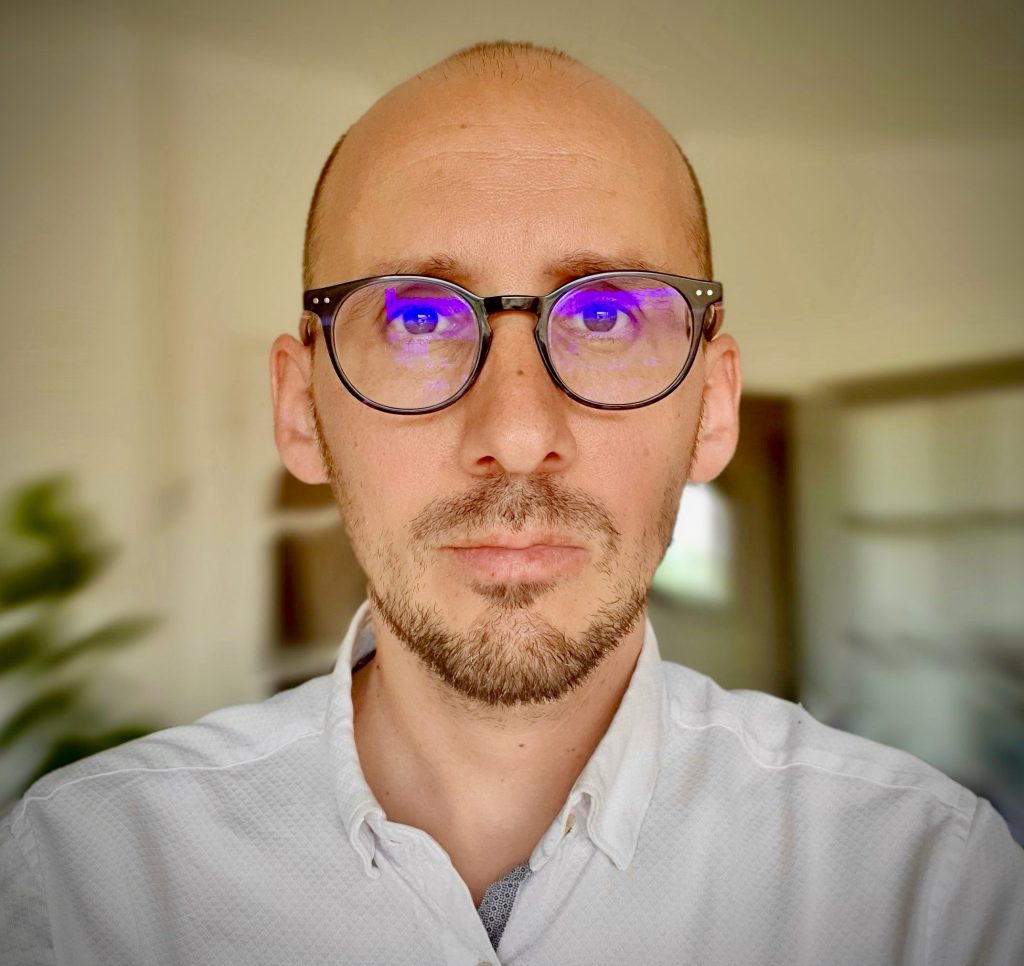 A photograph of Thomas Pierret looking directly at the camera with a serious expression. He wears glasses and a white shirt.