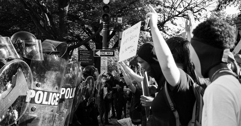 A black and white photograph of police in riot gear facing Black Lives Matter protestors