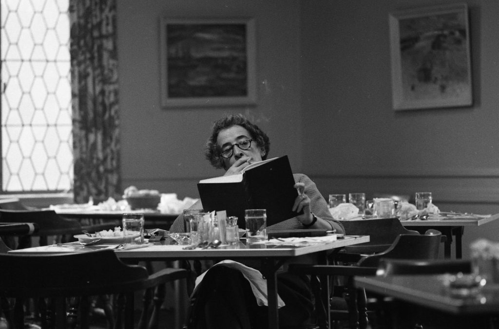 A black and white photograph of Hannah Arendt. She sits at a table set for a meal, reading a book and smoking a cigarette.