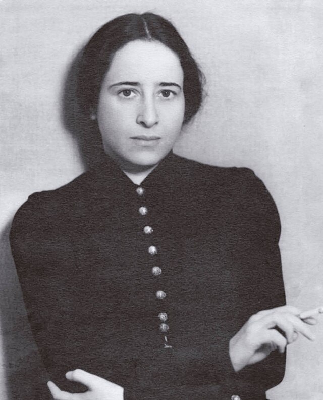A black and white photograph of Hannah Arendt at the age of 27. She wears a black dress and looks directly at the camera whilst holding a cigarette.
