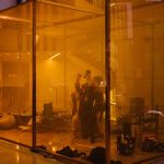 Photograph of an art installation in which multiple people crouch in an illuminated transparent box with their arms raised