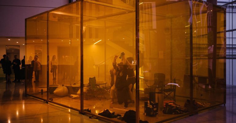Photograph of an art installation in which multiple people crouch in an illuminated transparent box with their arms raised