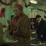 A blonde woman, dressed in a fur coat and red hat, stands at a shopping counter with a cigarette in her mouth, before a brunette woman wearing a Santa hat