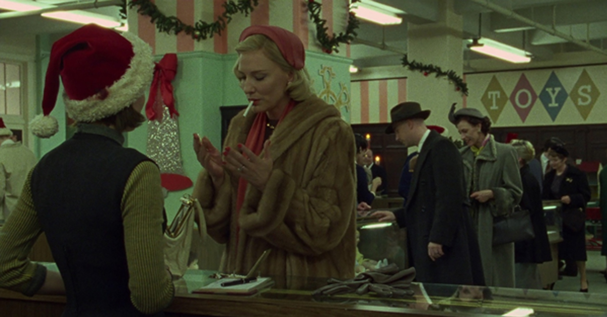 A blonde woman, dressed in a fur coat and red hat, stands at a shopping counter with a cigarette in her mouth, before a brunette woman wearing a Santa hat