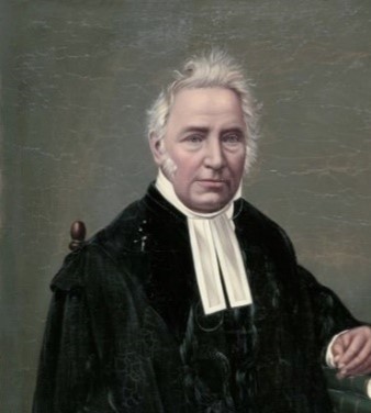 Colour portrait of a man with short white hair. He is looking straight ahead, wearing a black jacket with a white collar.