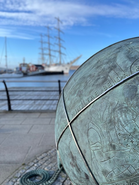 Close up image of a large blueish buoy, engraved with images of people preparing fish. In the background there is a sailboat sitting in water.