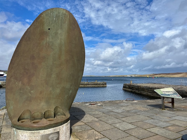 A metal statue of possibly a mussel shell, at a harbour with a bay in the background.