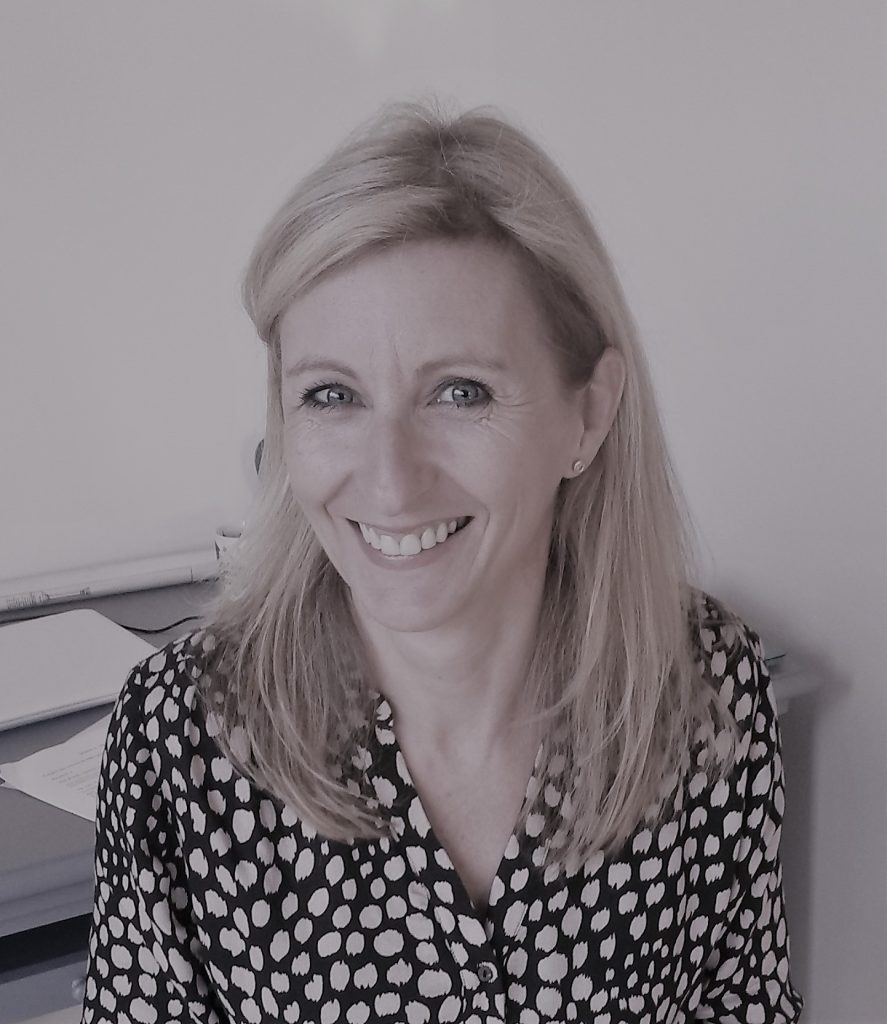 Portrait of Véronique Molinari. Véronique has shoulder-length blond hair and is looking straight ahead, smiling. She is wearing a black and white spotted shirt.