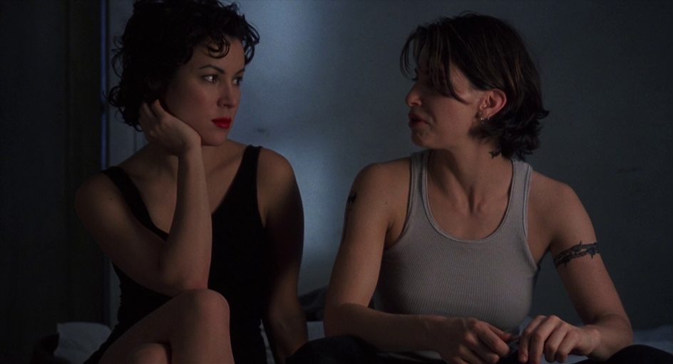 Two women, each wearing a tank top, sit upon a bed looking at each other