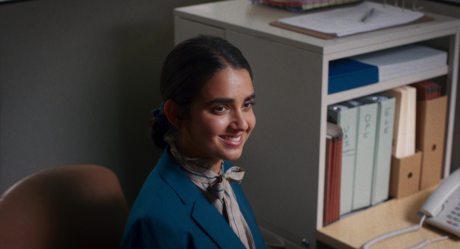 A young woman sits at a desk wearing a blue jacket and a neck tie smiling
