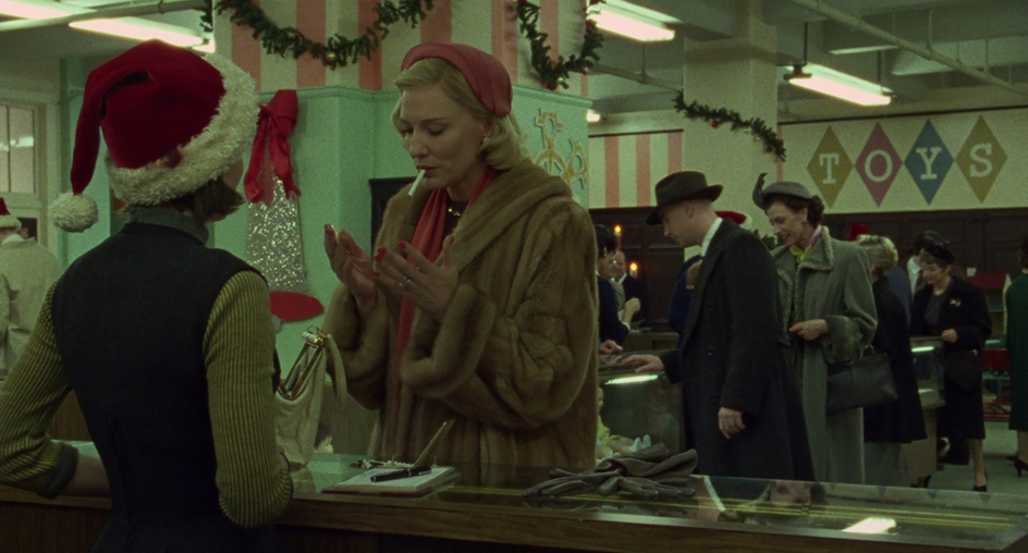 A blonde woman, dressed in a fur coat and red hat, stands at a shopping counter with a cigarette in her mouth, before a brunette woman wearing a Santa hat