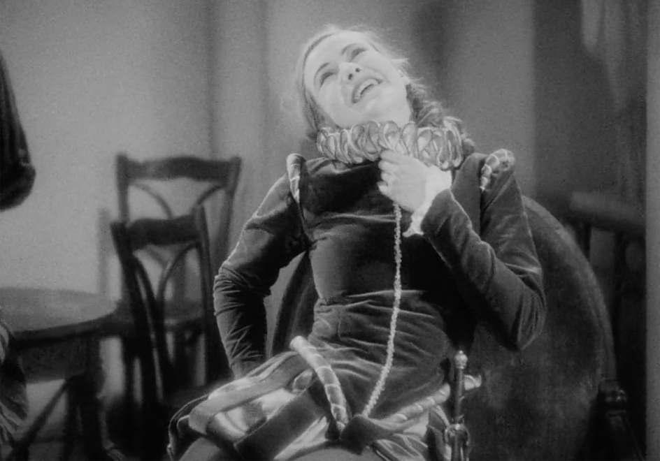 A black and white image of a woman dressed in male attire whilst laughing and clutching at her heart