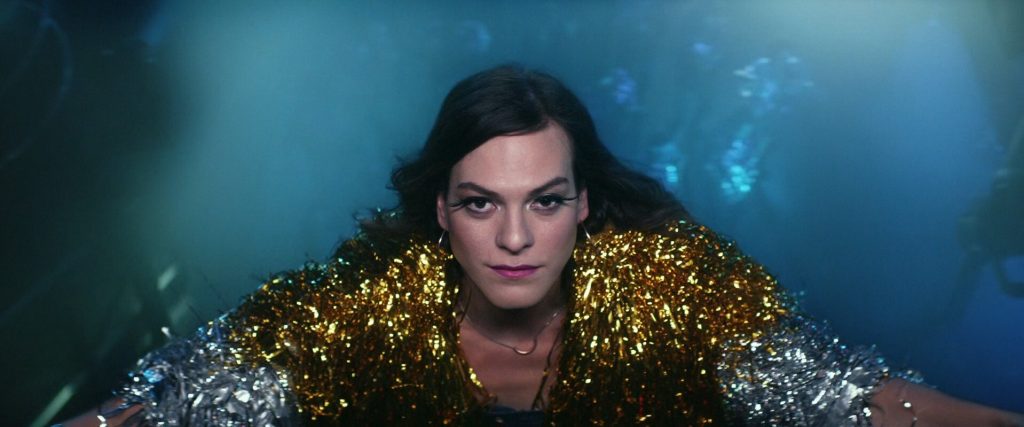A Latina woman with dramatic winged eyeliner wearing a gold and silver tinsel coat stares up at the camera, her arms outstretched against a murky blue backdrop.