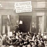 A crowd gathered outside a house, that features a banner above the door that reads 'No surrender. God help the sheriff who enters here.'