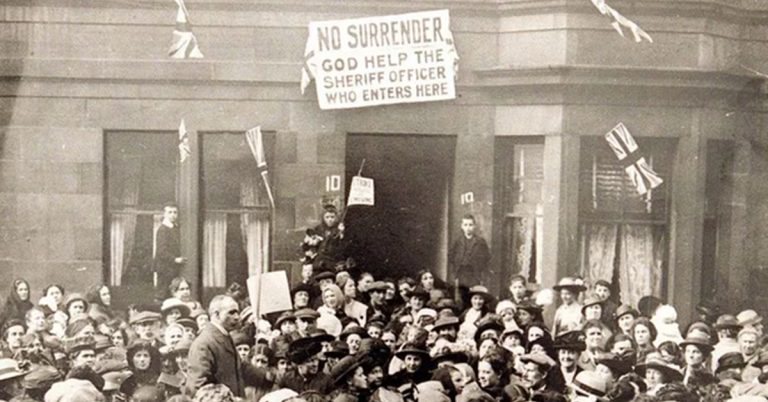 A crowd gathered outside a house, that features a banner above the door that reads 'No surrender. God help the sheriff who enters here.'
