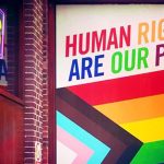 Progress flag emblazoned on office window with rainbow text which says, “Human Rights Are Our Pride.”