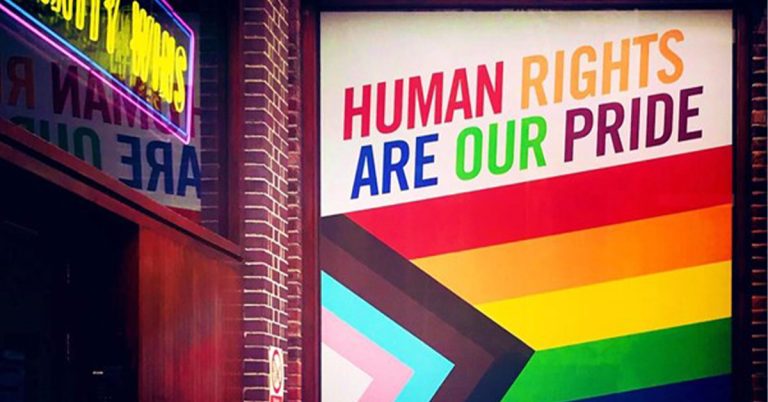 Progress flag emblazoned on office window with rainbow text which says, “Human Rights Are Our Pride.”