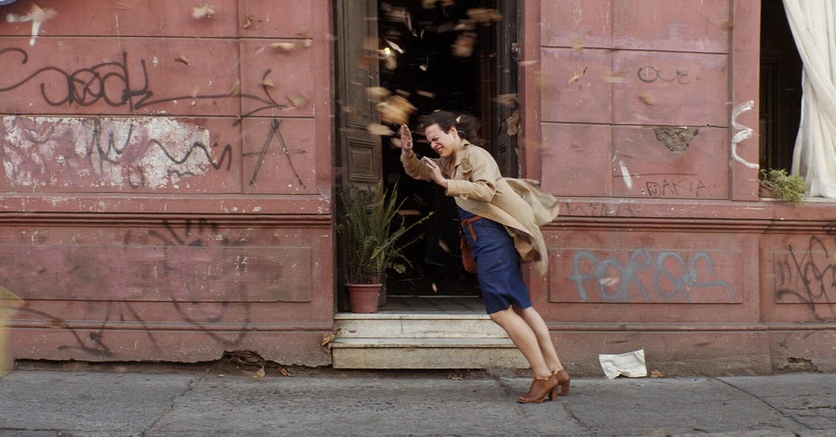 A woman leans forward at an impossible angle into the wind. She is framed by a dark doorway set in a pale red building covered in graffiti.
