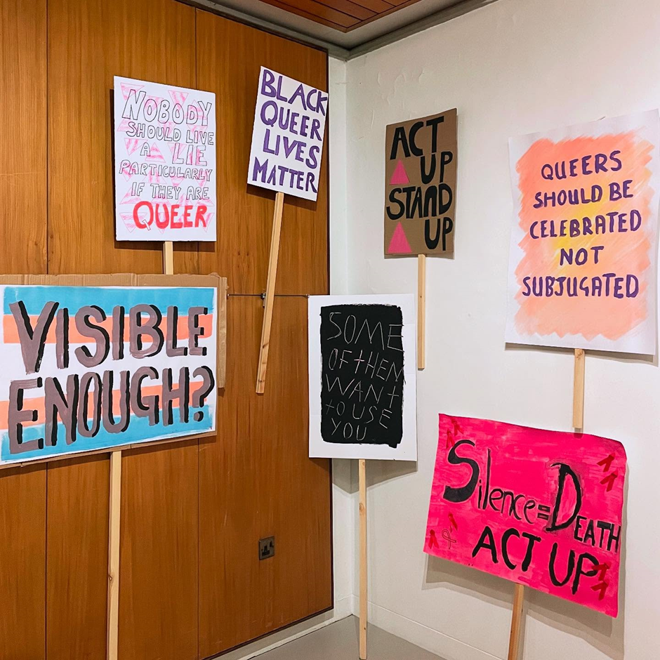 Collection of protest signs with various colours which say, “Visible Enough?”, “Nobody Should Live A Lie Particularly If They Are Queer,” “Black Queer Lives Matter,” “Act Up Stand Up,” “Some Of Them Want To Use You,” “Silence = Death ACT UP,” “Queers Should be Celebrated Not Subjugated.”