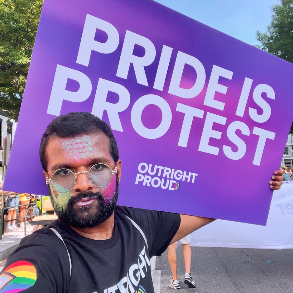 Man wearing various colours of glitter on face holds up purple sign on back which says, “Pride is Protest.”
