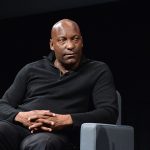 John Singleton sits on a grey chair, with his hands resting on his legs. He is facing towards the right of the frame, listening to someone who is out of frame.