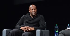 John Singleton sits on a grey chair, with his hands resting on his legs. He is facing towards the right of the frame, listening to someone who is out of frame.