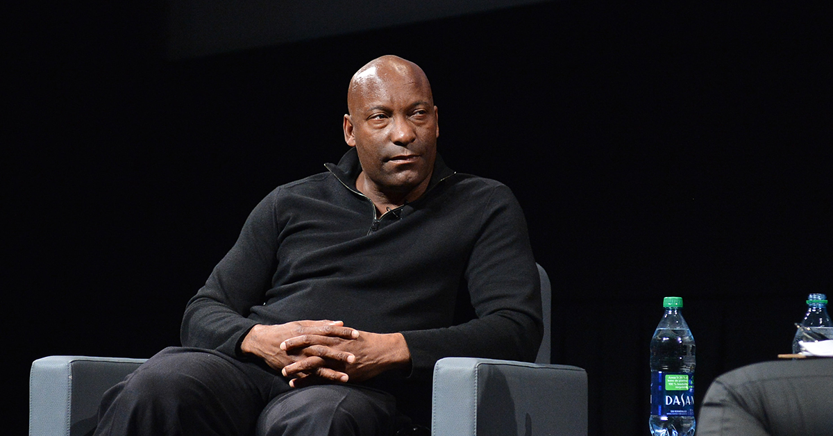 John Singleton sits on a grey chair, with his hands resting on his legs. He is facing towards the right of the frame, listening to someone who is out of frame.