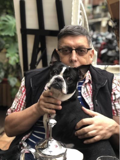 A man with glasses and salt-and-pepper hair sits facing the camera holding a black-and-white French bulldog.