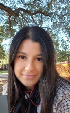 Headshot of a woman with long dark hair, wearing a checked jacket and a black scarf, with an outdoor background featuring trees.