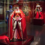 Exhibit display of Wonder Woman costumes in a museum. In the foreground, a mannequin wears a red and gold costume with a starry red cape inside a glass case. Behind it, another mannequin displays a blue suit, while to the right, a more modern Wonder Woman costume with armor, sword, and shield is showcased. A large poster of Wonder Woman is mounted on the wall in the background.