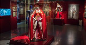 Exhibit display of Wonder Woman costumes in a museum. In the foreground, a mannequin wears a red and gold costume with a starry red cape inside a glass case. Behind it, another mannequin displays a blue suit, while to the right, a more modern Wonder Woman costume with armor, sword, and shield is showcased. A large poster of Wonder Woman is mounted on the wall in the background.