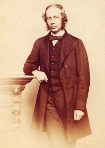 A man dressed in a Victorian-style suit, stands centre frame, his arm resting on a mantelpiece.