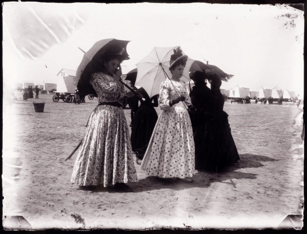 A group of five women on the beach holding parasols, with bathing machines in the background.