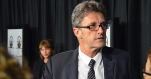 A man with salt-and-pepper hair stands in front of a black curtain, wearing a black and white tuxedo, looking off to the side