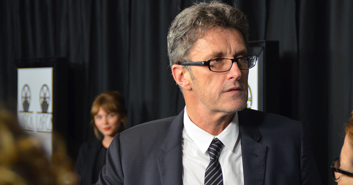A man with salt-and-pepper hair stands in front of a black curtain, wearing a black and white tuxedo, looking off to the side