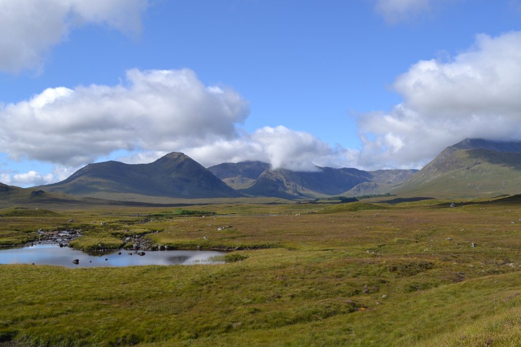 Landscape image of moorland with mountians and blue sky in the background. There is a small loch and stream in the left foreground.