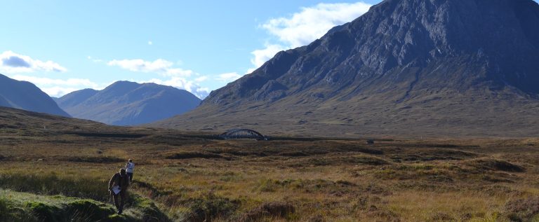 Landscape photograph of a heather-covered valley with mountains on either side. There is a small bridge in the middle-distance and two people holding clipboards and looking at the land in the foreground.