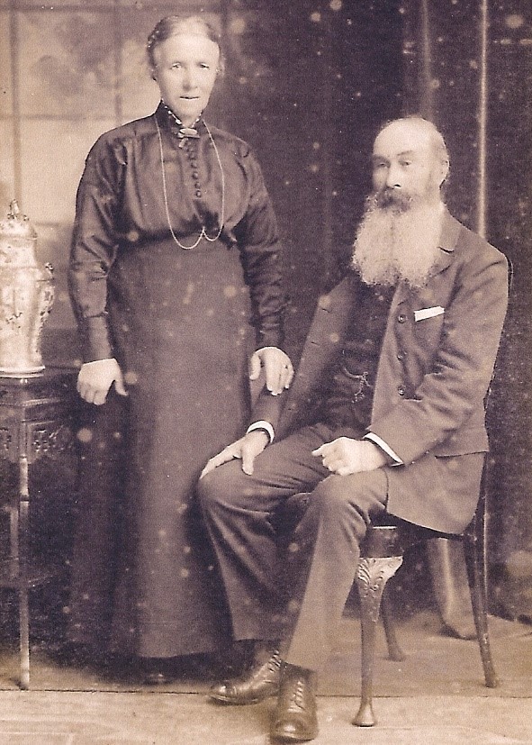 A sepia-toned studio portrait of an older couple. The woman stands beside a seated man with a long beard. Both wear formal Victorian-era clothing; she in a high-necked dark dress with a long chain necklace, and he in a three-piece suit. They pose beside a small ornate table with a decorative vase.