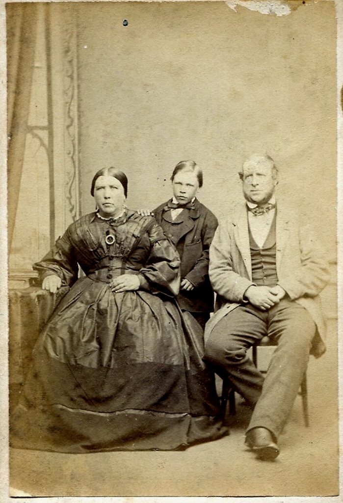 An early studio photograph showing a seated man and woman with a young boy standing between them. The woman wears a full, dark, crinoline-style dress with a brooch; the man wears a waistcoat and jacket. The boy is dressed formally in a suit. The background features a painted wall or curtain.