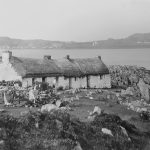 Black and white photograph of a traditional stone cottage with a thatched roof, set on a rocky coastal landscape overlooking the sea, with distant hills and scattered buildings across the water.