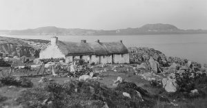 Black and white photograph of a traditional stone cottage with a thatched roof, set on a rocky coastal landscape overlooking the sea, with distant hills and scattered buildings across the water.