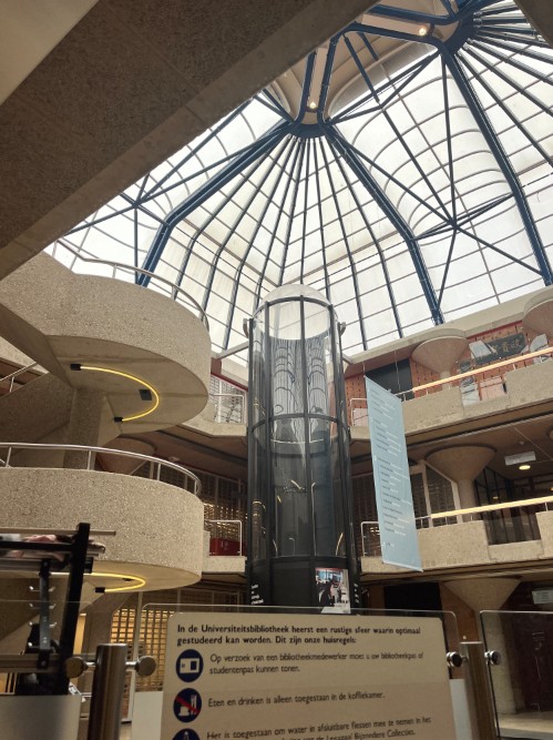 Interior of a large university library with a tall glass elevator in the center and multiple curved balconies surrounding it. The ceiling features a striking glass dome supported by blue metal beams, allowing natural light to fill the space. A sign in Dutch with library guidelines is visible in the foreground.