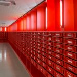 A brightly lit hallway lined with red numbered mailboxes or storage lockers on both sides. The walls, drawers, and lighting fixtures are all red, creating a striking monochromatic design. The polished floor reflects the light, and a white desk is visible on the left side near the entrance.