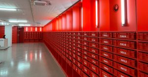 A brightly lit hallway lined with red numbered mailboxes or storage lockers on both sides. The walls, drawers, and lighting fixtures are all red, creating a striking monochromatic design. The polished floor reflects the light, and a white desk is visible on the left side near the entrance.