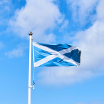The Scottish flag blows in the wind against a blue sky, with light white clouds
